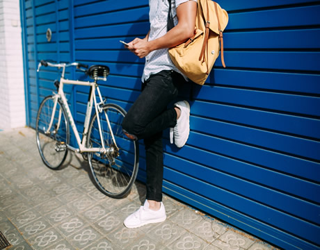 Male student with backpack and bike leaning against blue garage door looking at a cell phone